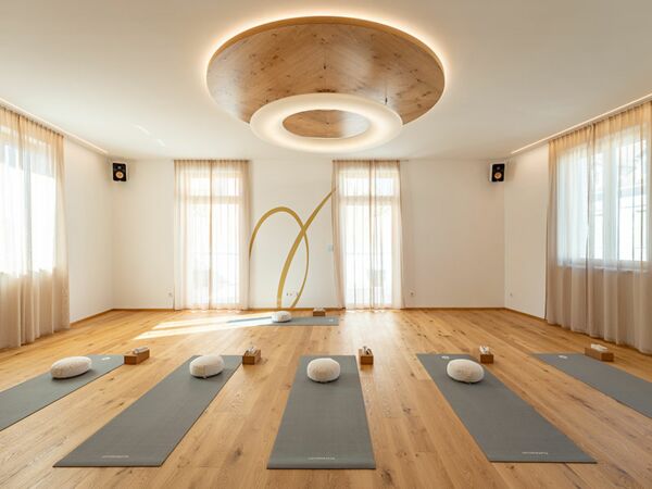 Der Yoga Raum ist hell und freundlich, es liegen graue Matten am Eichen Parkett Boden und weiße Sitzkissen auf den Matten.