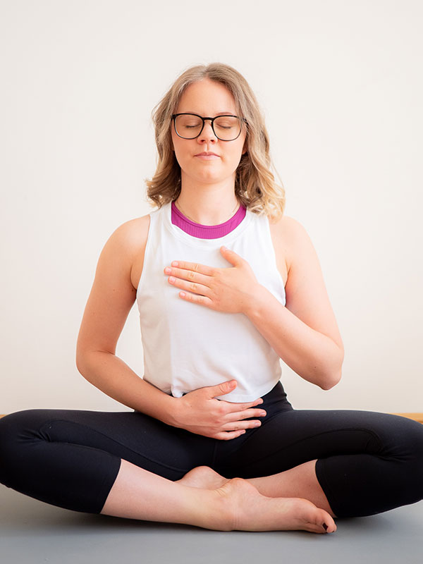 Hanna sitzt im Schneidersitz auf der grauen Yoga Matte, hat einen Hand am Bauch und die andere Hand am Herzen. Ihre Augen sind geschlossen und sie lächelt sanft.