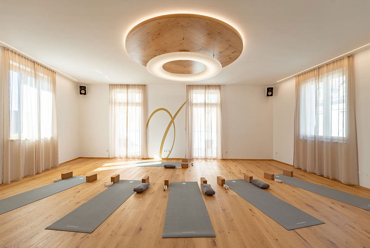 Der Yoga- & Workshop-Raum hat einen Eichen-Parkett Boden, mehrere Fenster und ein Dachterrasse. Am Boden liegen graue Yoga Matten mit Blöcken und kleinen Kissen. Er ist sehr hell und freundlich.